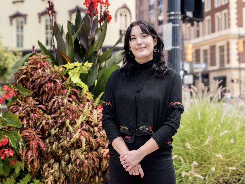 A person with dark hair stands outdoors in front of vibrant plants. Theyre wearing a black outfit with floral details, smiling slightly. In the background, there are city buildings and a street with traffic lights.