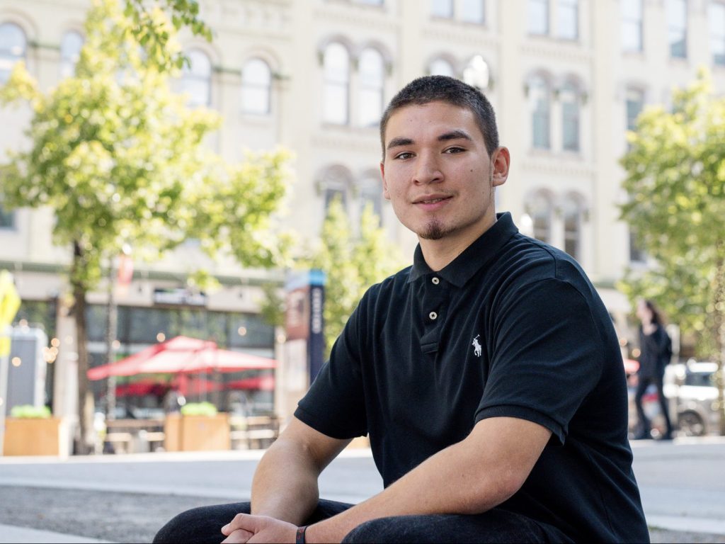 A young person wearing a black polo shirt sits outdoors in an urban setting with trees and a building in the background. The person is looking at the camera and smiling slightly.