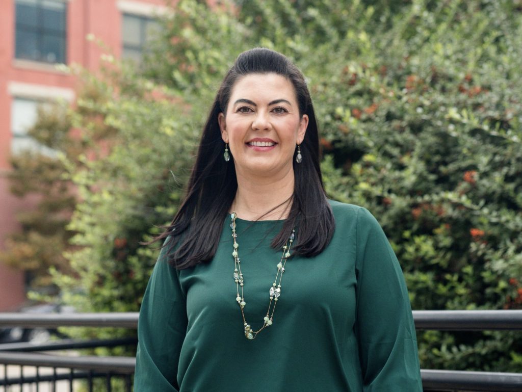 A woman with long dark hair, wearing a green blouse and necklace, stands smiling in front of greenery and a brick building.