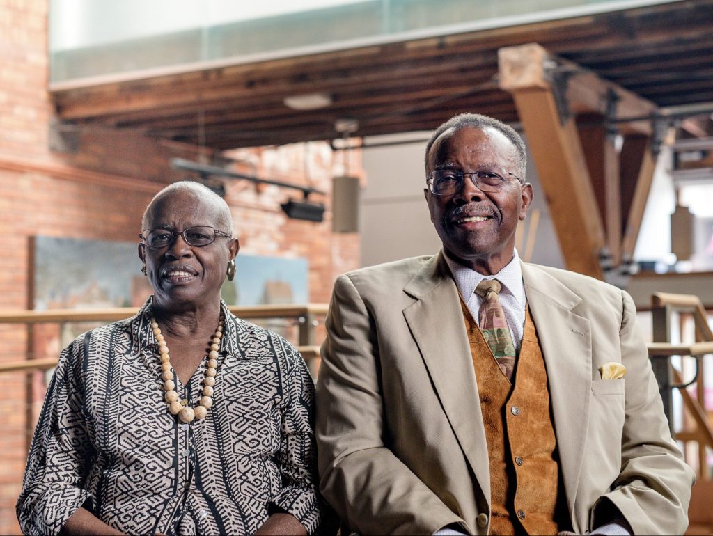 An elderly couple is seated, smiling at the camera. The woman wears a patterned dress and beaded necklace, while the man wears a light suit with a tie and vest. They are indoors, with a backdrop of brick walls and wooden beams.