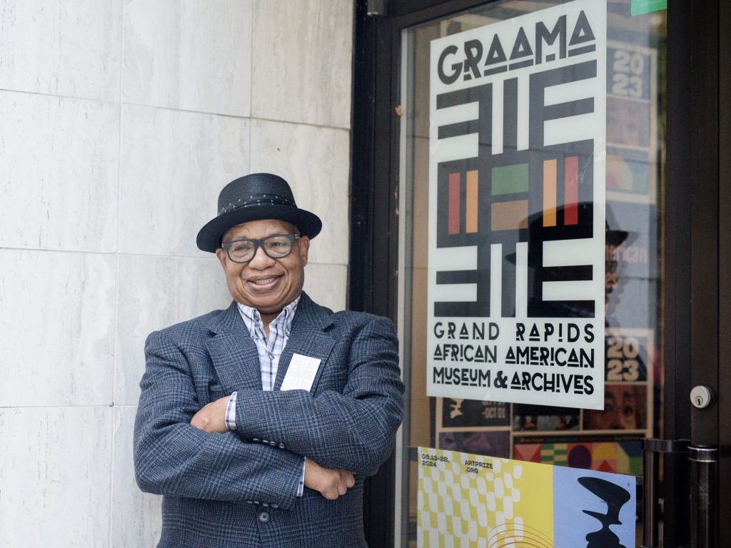 A person wearing a plaid jacket and black hat stands with arms crossed in front of a door. The door has a sign for the Grand Rapids African American Museum & Archives with an abstract design.