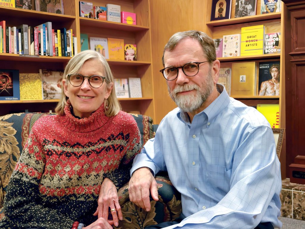 A woman in a red patterned sweater and a man in a blue shirt sit together, smiling, in a cozy library filled with books on the shelves behind them.