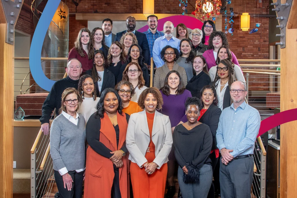 A diverse group of 29 people, including men and women of various ages, pose together on a staircase inside a building. The background includes a wooden banister, brick wall, and colorful hanging decorations.