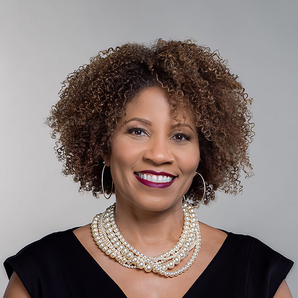 A person with curly hair smiles, wearing a black top, hoop earrings, and a layered pearl necklace. The background is gray.