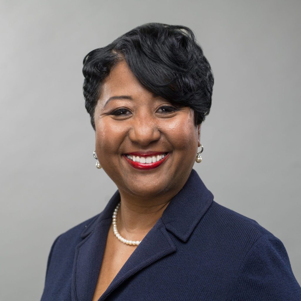 A smiling woman with short black hair wears a pearl necklace and earrings, along with a dark blazer, against a plain gray background.