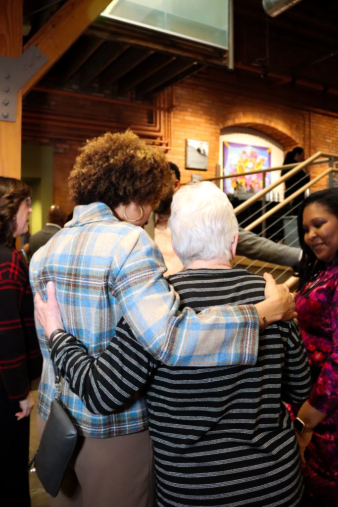 Two women seen from the back, one with curly hair in a plaid jacket and the other with short white hair in a striped top, embrace while standing in an art gallery. Other people and a staircase are visible in the background.
