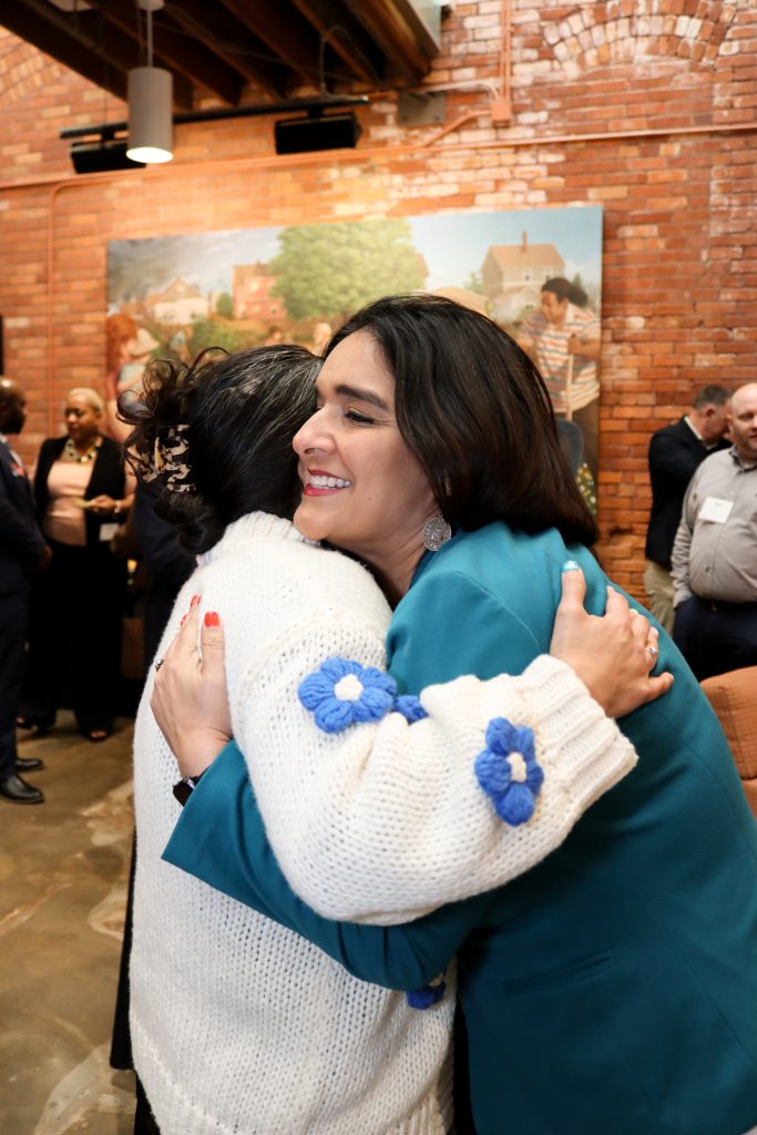 Two women warmly hugging in a room with people in the background. One woman wears a white sweater with blue flowers, the other wears a teal jacket. A large painting and exposed brick wall are visible in the background.