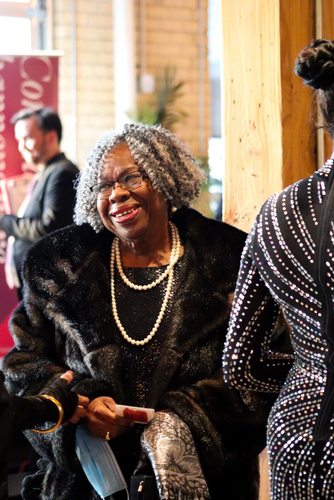 An elderly woman with curly gray hair is smiling while wearing a black fur coat and pearl necklace. She is interacting with a person in a sparkly dress in a warmly lit room. A blurred figure is in the background.