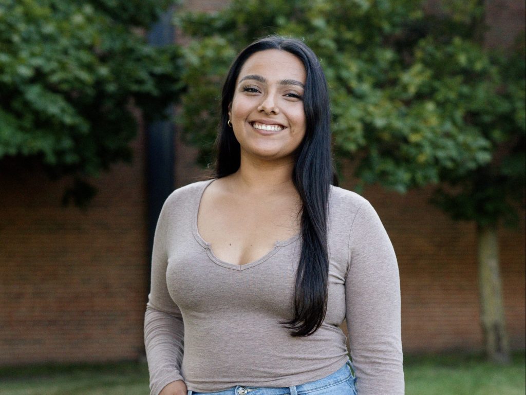 A person with long dark hair smiles while standing outdoors. They wear a long-sleeve beige top and light blue jeans. Green trees and a brick wall are in the background.